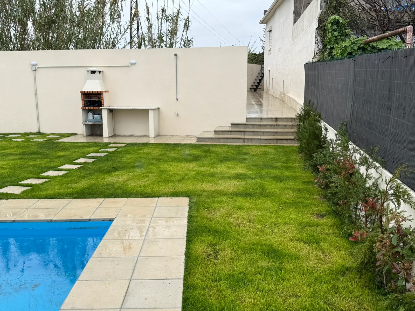 Jardim com relva, piscina parcialmente visível, barbecue e acesso lateral